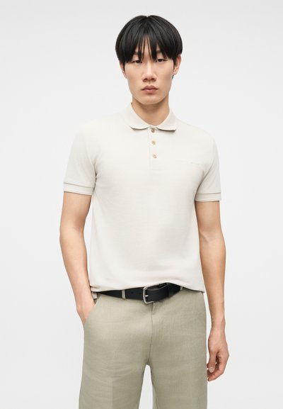 Νεαρός άνδρας με μαύρα μαλλιά, που φοράει ανοιχτό μπεζ πόλο πουκάμισο τυλιγμένο μέσα σε ανοιχτό λαδί παντελόνι με μαύρη ζώνη, τα χέρια στις τσέπες, ουδέτερο φόντο.