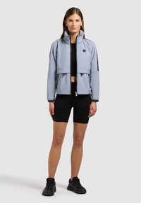Jeune femme debout, portant une veste zippée bleu clair, un crop top noir, un short cycliste noir et des chaussures de sport noires, sur un fond clair uni.