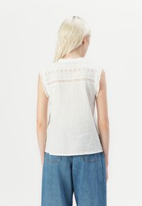 Weiße ärmellose Bluse mit strukturierten Polka-Punkten, Spitzenakzenten auf der Rückseite und einem gerüschten Ausschnitt. Kombiniert mit blauen Jeans.