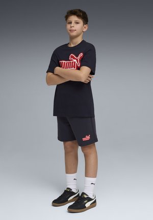 Niño de pie con los brazos cruzados, vistiendo una camiseta negra de Puma, pantalones cortos a juego, calcetines blancos de Puma y zapatillas negras de Puma sobre un fondo gris.