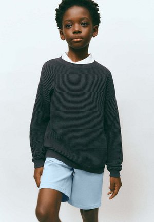 Junges Kind mit kurzen lockigen Haaren, das einen dunklen Rippsweater über einem weißen Hemd und hellblauen Shorts trägt, vor einem einfarbigen Hintergrund stehend.