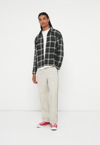 Camicia di flanella a quadri bianco e nero, t-shirt a girocollo bianca, pantaloni beige con strisce laterali e sneakers rosse.