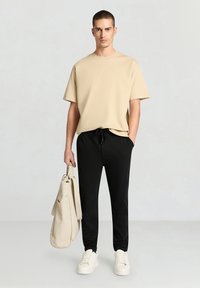 Beiges T-Shirt mit kurzen Ärmeln, kombiniert mit schwarzen Jogginghosen. Das Modell hält eine cremefarbene Tragetasche. Weiße Turnschuhe vervollständigen das Outfit. Glatter, lässiger Stil.