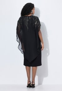Femme avec des cheveux bouclés portant une robe midi noire et un châle noir transparent avec des détails en perles argentées, se tenant contre un mur blanc, de dos.