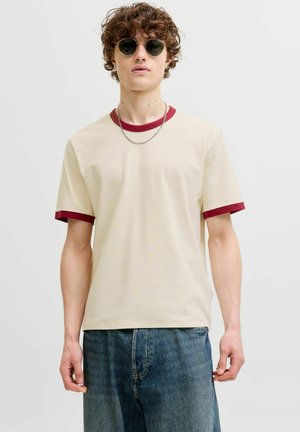 Jeune homme aux cheveux bouclés porte des lunettes de soleil rondes, un t-shirt beige avec un liseré rouge au col et aux manches, et un jean bleu sur un fond uni.