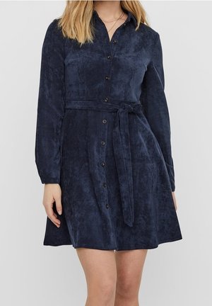 Granatowa sukienka z korduroju z długimi rękawami, z przodu zapinana na guziki i z pasującym paskiem z materiału. Posiada gładką fakturę i rozkloszowany dół.