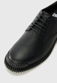 Zapato negro de cuero con cordones, puntera texturizada y suela de goma gruesa y ranurada sobre un fondo claro.