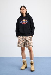 Black hoodie with "Dickies" logo, patterned beige shorts with animal prints, and tan lace-up boots. Plain background and blue floor.