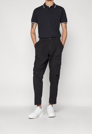 Polo shirt bleu marine avec des accents de col blanc, associé à un pantalon cargo noir avec des poches latérales et des baskets blanches. Style décontracté.