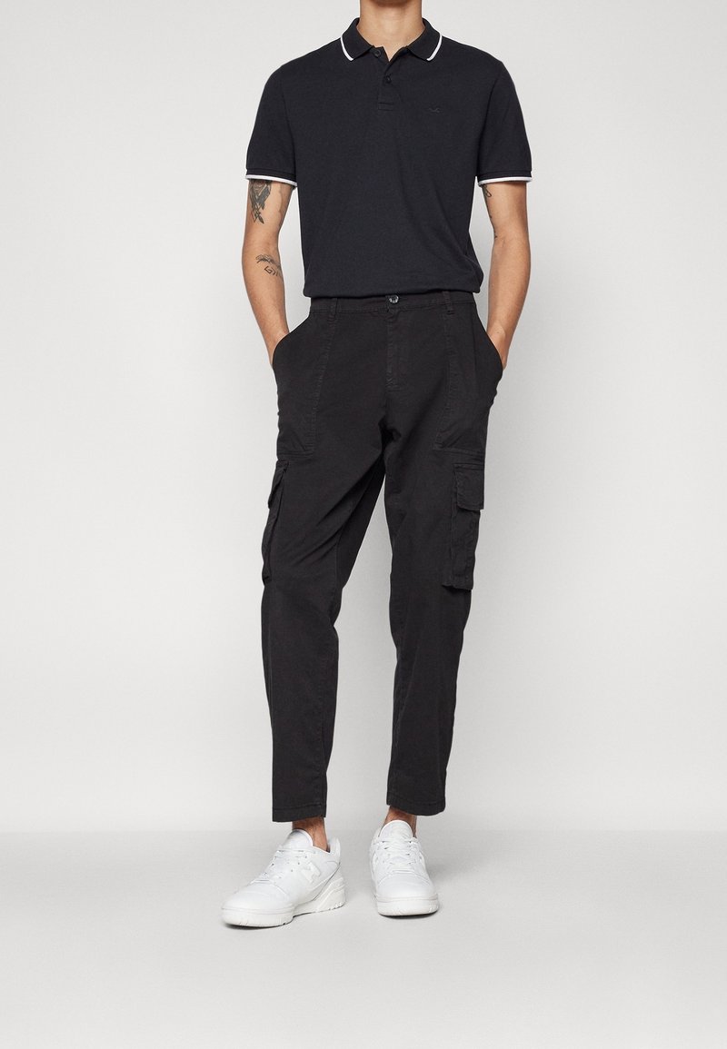 Polo shirt bleu marine avec des accents de col blanc, associé à un pantalon cargo noir avec des poches latérales et des baskets blanches. Style décontracté.