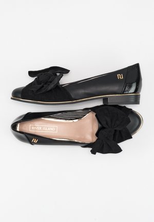 Para czarnych płaskich butów z dużymi materiałowymi kokardami na palcach, złotymi akcentami i etykietą "River Island" wewnątrz jednej z butów.