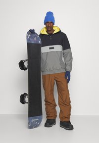Tabla de snowboard con parte superior oscura, acentos azules y fijaciones; modelo con chaqueta gris y amarilla, pantalones marrones, gorro azul y guantes.