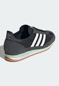 Svarta sportskor med övre i tyg och mocka, tre vita ränder, grön mellansula, gummisulor och en texturerad detalj vid hälen.