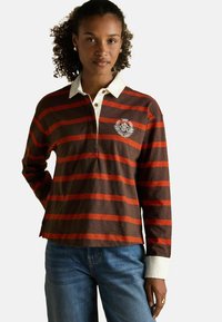 Frau mit lockigem Haar trägt ein braun-orangestreifiges Rugby-Shirt mit weißem Kragen und Manschetten, kombiniert mit blauen Jeans, steht mit einer Hand nach hinten.