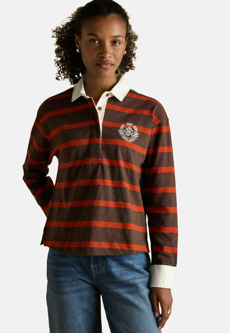 Frau mit lockigem Haar trägt ein braun-orangestreifiges Rugby-Shirt mit weißem Kragen und Manschetten, kombiniert mit blauen Jeans, steht mit einer Hand nach hinten.