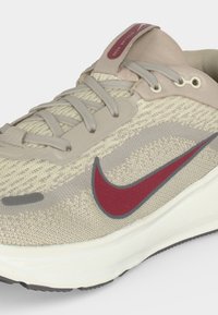Μπεζ και γκρι παπούτσι τρεξίματος Nike με κόκκινο λογότυπο Swoosh, γκρι κορδόνια, πάνω μέρος από πλεκτό ύφασμα και λευκή σόλα, εμφανιζόμενο από πλάγια γωνία.