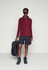 Bordeauxkleurige fleece pullover met een halve rits, zwarte cargoshorts, gecombineerd met zwarte wandelschoenen. Model houdt een zwarte rugzak vast.