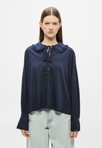 Μπλούζα σε ναυτικό μπλε με γιακά, φραμπαλάδες στον γιακά, κλείσιμο με κορδόνι και μακριά μανίκια με χρυσές λεπτομέρειες στα μανσέτες. Λείο ύφασμα.