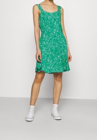 Grünes Blumenkleid mit quadratischem Ausschnitt, breiten Trägern und ausgestelltem Saum. Kombiniert mit weißen High-Top-Sneakern. Weicher, gemusterter Stoff.