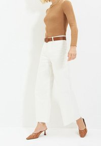 Haut marron à manches longues côtelé associé à un pantalon blanc à jambes larges, maintenu par une ceinture marron. Des mules marron à bouts pointus complètent la tenue.