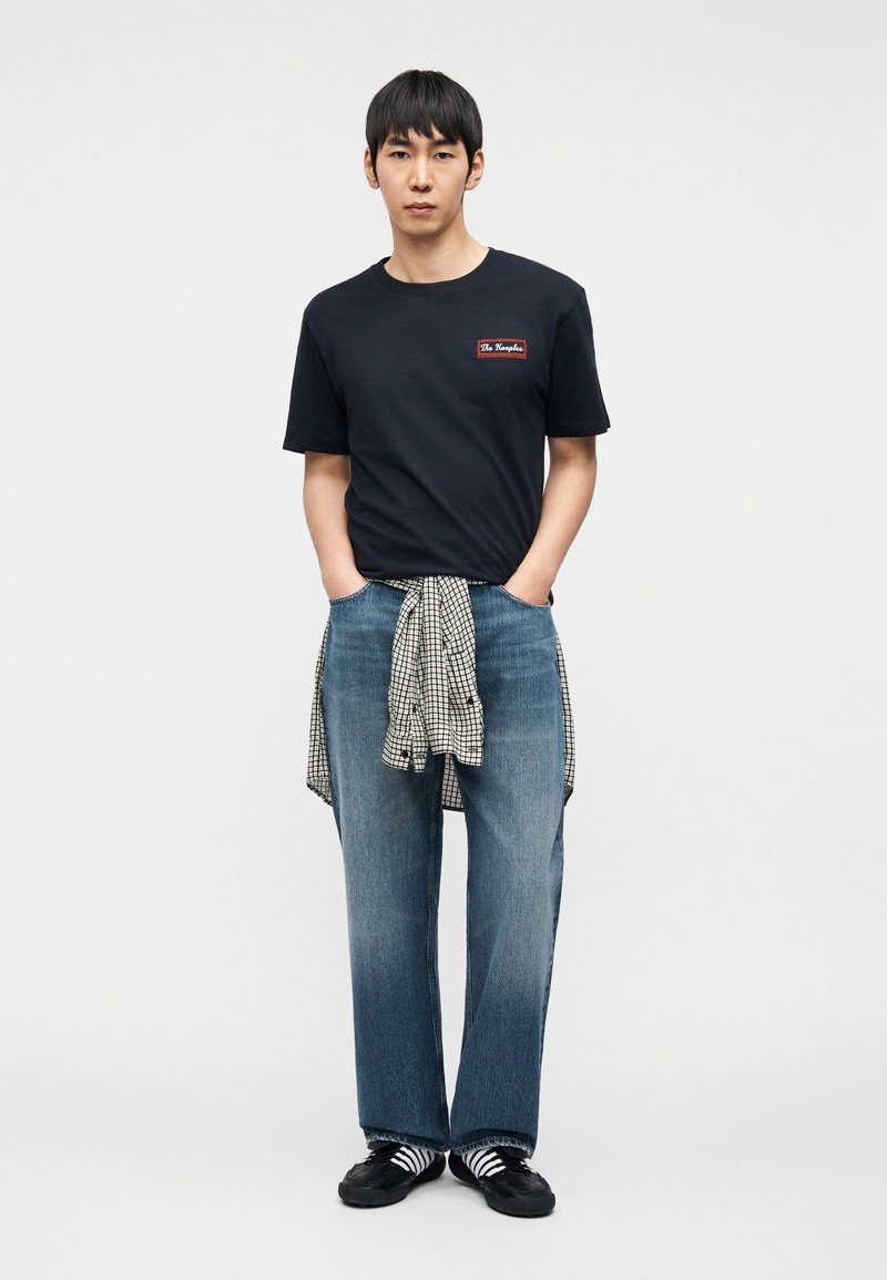 Navy cotton t-shirt with logo patch, light blue wide jeans, black and white striped socks, and black shoes with checkered shirt tied at the waist.