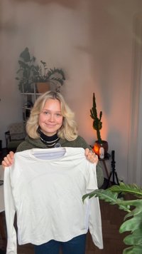 Wit sportshirt met lange mouwen, lichtgewicht, met een gestructureerde voorkant en een afgeronde zoom; vastgehouden door een persoon tegen een neutrale achtergrond.