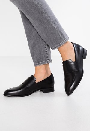 Des chaussures noires en cuir à enfiler avec un petit talon, dotées d'une finition lisse et d'une large bandoulière au niveau du métatarse. Portées avec un jeans gris cropped.