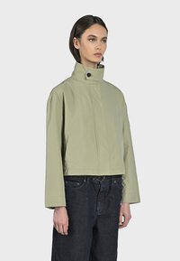 Beskarvad, ljusgrön jacka med hög krage, framknäppning, sidofickor och vida ärmar. Tyget verkar slätt och lätt.