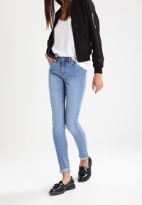 Black bomber jacket, white top, light blue skinny jeans, and black loafers. Features include zippered pockets and rolled cuffs.