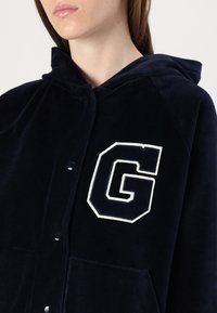 Mornarplavi baršunasti dukserica s prednjim gumbima, dva džepa i bijelim izvezenim logom "G" s lijeve strane. Glatka tekstura, opušteni kroj.