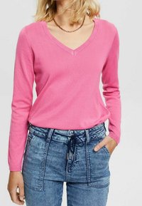 Różowy sweter z dekoltem w serek wykonany z miękkiego materiału z długimi rękawami. Do tego wysokie niebieskie dżinsy z bawełny z marszczonym paskiem ze sznurkiem i bocznymi kieszeniami.