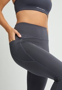 Dunkelgraue Leggings aus dehnbarem Stoff, mit hohem Bund und Seitentasche. Glatte Textur mit Flachnähten für zusätzlichen Komfort.