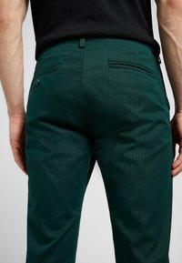 Grüne Hosen aus einem glatten, strukturierten Stoff mit feinen Streifenmustern. Sie verfügen über Gesäßtaschen und eine taillierte Passform.