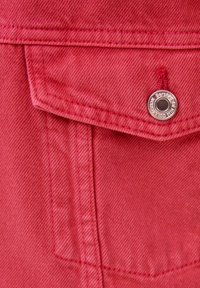 Tissu en denim rouge avec une poche poitrine, comportant un bouton métallique avec un logo gravé. La texture est en twill avec des coutures visibles.