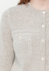 Lys grå cardigan laget av mykt strikket stoff, med ribbestrikket krage, fem knapper, og en enkelt brystlomme med knapp.