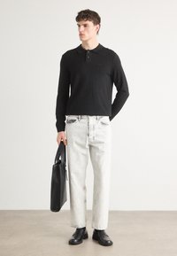 Melna garām piedurknēm polo krekls ar apkakli, saskaņots ar gaišām, nolietotām džinsām. Modelis tur melnu somu un valkā melnus mokasīnus.
