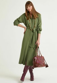 Olivgrünes Wickelkleid mit langen Ärmeln und Gürtel, kombiniert mit einer strukturierten burgunderroten Handtasche und kniehohen gesteppte Stiefeln.