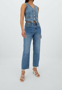 Kvinna i ärmlös knäppt denimväst, högmidjade blå jeans med råa kanter, och vita sandaletter med klack mot en enhetlig bakgrund.