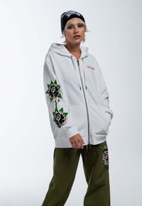 Sudadera blanca con cremallera y detalles bordados de flores en las mangas, que cuenta con capucha y bolsillos tipo canguro. Combinada con pantalones verdes oliva.