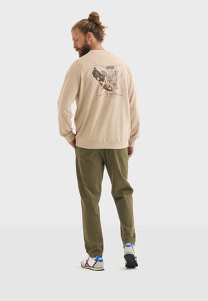 Beige sweatshirt met ronde hals en grafische print op de achterkant, gecombineerd met olijfgroene broek en witte sneakers met blauwe accenten.