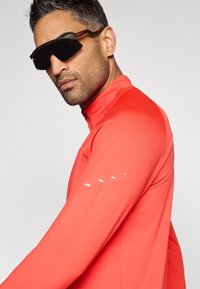 Κόκκινο μακρυμάνικο τοπ με ψηλό γιακά και διακριτικές ανακλαστικές ρίγες στο μανίκι, συνδυασμένο με μαύρα γυαλιά ηλίου με έντονο σχέδιο.