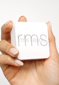 Compact carré blanc aux coins arrondis, avec le texte "rms beauty" dans une police brune minimaliste, tenu entre des doigts avec un vernis à ongles léger.