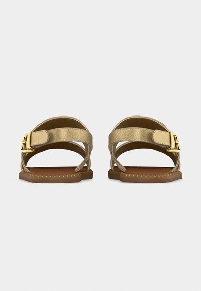 Φλατ χρυσά σανδάλια με λουράκια στον αστράγαλο, κλεισίματα με χρυσή αγκράφα και καφέ σόλες, εμφανιζόμενα από πίσω σε λευκό φόντο.