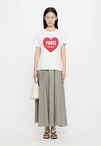 Valkoinen puuvillapaita, jossa on punainen sydänkuvio ja teksti "PARIS L'amour". Yhdistetty pitkän raidallisen, laskostetun harmaan hameen ja mustien hihnallisten sandaalien kanssa.
