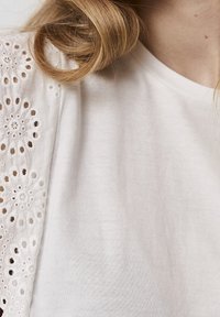 White cotton shirt with an intricate lace overlay on the left sleeve featuring floral cutouts. Soft texture and relaxed fit.