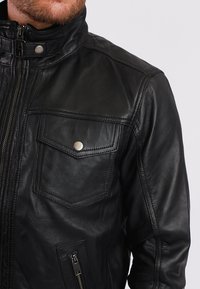 Schwarze Lederjacke mit hohem Kragen, Reißverschluss und zwei Vorderentaschen. Glatte Textur, minimalistisches Design und silberne Hardware-Akzente.