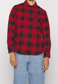 Camisa de cuadros en rojo y negro con botones, de manga larga, con un bolsillo en el pecho y botones negros, combinada con jeans de denim azul claro.