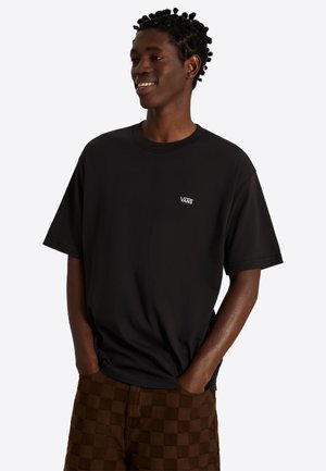 Jeune homme aux cheveux tressés souriant, portant un t-shirt noir Vans et un short marron à carreaux, mains dans les poches, fond blanc.