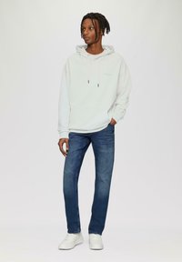 Jasnoszara bluza z kapturem, z trokami i kieszenią z przodu, połączona z ciemnoniebieskimi dżinsami i białymi sneakersami. Prosty design, brak widocznych wzorów.