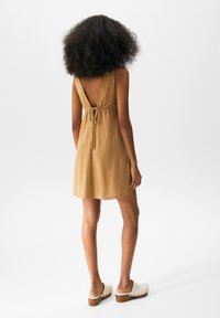 Femme aux cheveux bouclés portant une mini-robes sans manches couleur beige avec un dos ouvert et des sabots blancs à plateforme, debout sur un fond blanc.